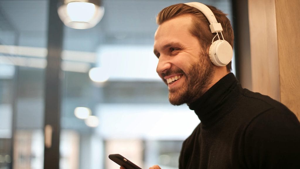 A man in profile smiles while wearing headphones and holding his mobile phone. Podcasts are a great way to improve your English!