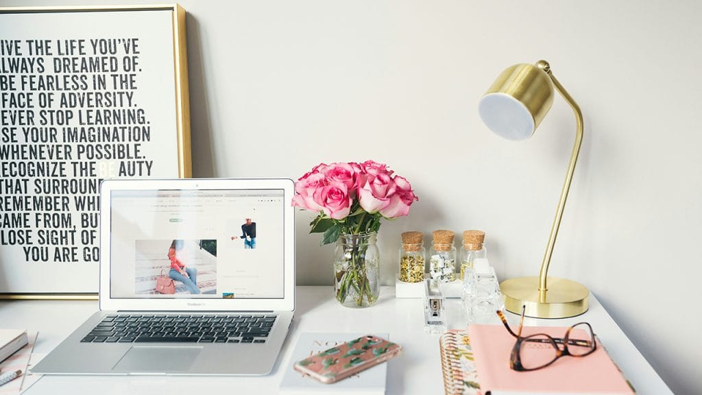 A bright learning space in a white room, a desk with an inspirational quote, a laptop, flowers, a lamp, notebooks, a book, glasses, a mobile phone and various decorations. A great place to keep you sharp to learn English fast.