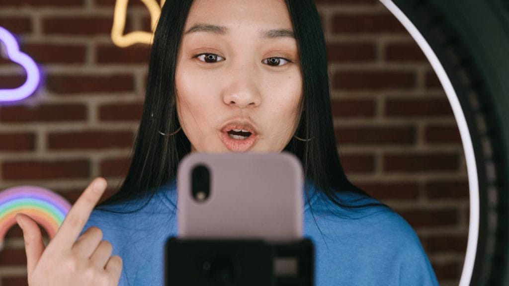 A young woman practicing speaking alone by recording a video on her smartphone in front of a ring light and neon signs.